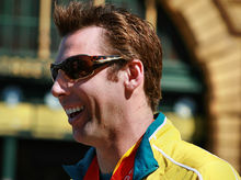 Profile of a smiling, brown haired young main, wearing sunglasses, wearing a yellow jacket tracksuit with a green collar, with a red medal ribbon around his neck.