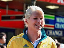 Young woman with bleached blonde hair, short and tied up, smiles. She is wearing a yellow tracksuit with a green collar, with the logo of Adidas and the coat of arms of Australia visible.