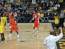  a man in a white shirt makes a signal to basketball players on the court with his fist in the air from the sidelines. He is viewed from behind.