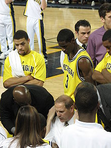 basketball players in maize uniforms have their attention on a man in a white shirt who is seated or kneeling below them.  They look over his shoulders as other people look on.