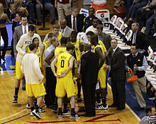 The basketball players standing in maize uniforms and men in suits are huddled around a man in a white shirt and dark pants.