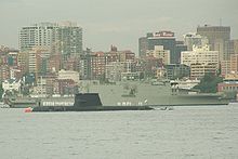 A submarine sitting in calm water, in front of a large warship, and with numerous tall buildings in the background. White uniformed personnel are standing on the decks of both vessels.