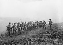 A line of soldiers in battle equipment face another soldier who is addressing them on a gentle slope. Behind them smoke or fog obscures the rest of the terrain