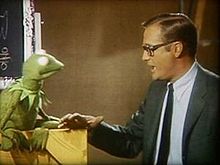Man on the right, wearing a conservative suit and tie, speaking to the Muppet Kermit on the left.