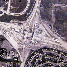 A bird's-eye view of a large highway interchange under construction. Several bridges are complete, but nothing is paved, aside from one highway crossing horizontally, which detours between the bridges.
