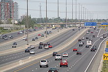 "A view of a wide freeway from the side. The freeway is divided into four segments, each of which contains three lanes (up to five in some places). The vehicles within the two sections nearest the camera are travelling away into the distance, whereas the vehicles within two sections further from the camera are approaching. Several signs dot the right side of the freeway, and two overhead gantries (also holding signs) are also visible."