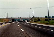 Driving down a six lane highway during the day. In front is a concrete bridge. The highway curves to the right as it passes beneath the bridge.