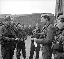 Officer with swagger stick, talking to six other men. In the background are other men and a mountain range