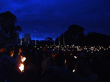 People gather with candles in the dim light of dawn in front of a large stone building and a line of flag poles
