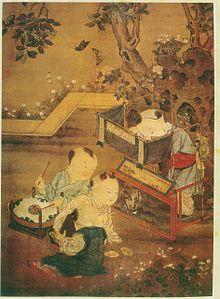 Three young boys sit and watch as a fourth boy dangles puppets from behind a small booth set up in a garden.