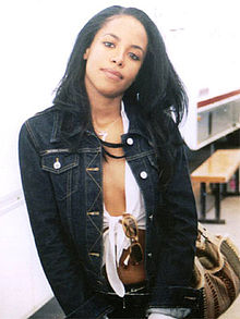 Head and upper torso of a young woman with long, black hair and a broad facial expression, wearing an unbuttoned navy blue jacket and a tank top underneath. Glasses hang from the center of her tank top and she holds a brown purse on the left side of her body.