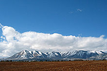 Snow covered mountains protruding from a plain with tilled soil in the foreground.
