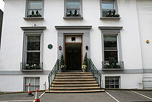 A flight of stone steps leads from an asphalt car park up to the main entrance of a white two-story building.  The ground floor has two sash windows, the first floor has three shorter sash windows.  Two more windows are visible at basement level.  The decorative stonework around the doors and windows is painted grey.