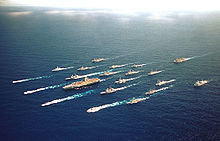 Aerial photograph of twenty-one ships, including an aircraft carrier, sailing in close formation.