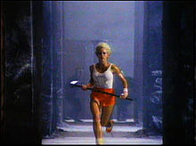 A young woman carrying a sledge hammer and wearing a white tank top with a drawing of a Macintosh runs from black figures in the background.