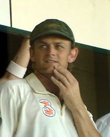 A man in a white cricket shirt and a baggy green cap, with his left hand on his chin, looking to his right