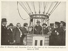 Two men wearing hats in the basekt of a balloon, surrounded by a group of men in suits