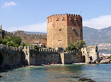 A eight-sided medieval tower built of red and yellow brick rises above a green sea in which swimmers play.  Stone walls run along the shore and further up from the tower.