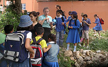 Several children dressed in blue wearing backpacks crowd around a small rock enclosure.