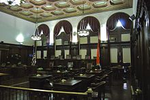 A large room with four arched windows, a bar, and multiple seats for legislators. Walls are wood paneled and the room is lit by two large chandeliers.