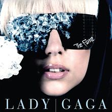 The face of a blond woman. She is wearing black glasses. The right side of the glasses is covered by blue crystals. On the bottom of the left side of the glasses, the word "The Fame" is written in white.