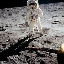An astronaut in an American Apollo-program spacesuit, standing on the flat, heavily footprinted landing area, with the utterly black sky of space above the horizon.