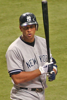 A Hispanic man walking while shouting at someone out of the picture. His helmet is emblazoned with a white "N" and "Y" intertwined, and "NEW YORK" is stitched in black letters across his button-down jersey. The player is holding a black baseball bat almost vertically with black, gray, and white gloves.