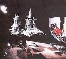 In a film studio, a director operates a crane-mounted camera aimed at a large grey model spacecraft, raised several feet in the air by a support structure underneath. The model is lit from one side by bright studio lights and has tall spires with many pieces protruding from them.