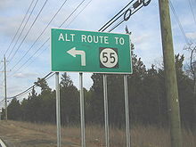 A green sign along a road lined with trees and power lines reading alt route to Route 55 with an arrow pointing to the left
