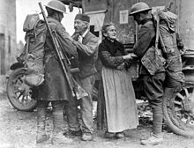 Two soldiers are warmly greeted by civilians - and elderly woman and man. A parked ambulance is behind them