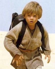 A blonde boy wearing a gray robe and a black backpack walks in a desert.