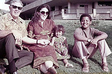 A young boy possibly in his early teens, a younger girl (about age 5), a grown woman and an elderly man, sit on a lawn wearing contemporary circa-1970 attire. The adults wear sunglasses and the boy wears sandals.