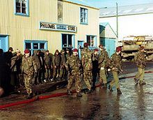 A group of armed soldiers in camouflage uniform with red berets guard a line of enemy soldiers, who stand in front of a large wooden building, which is painted yellow.  The road is wet, and the sky is blue.