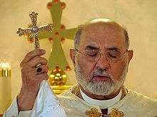 A middle-aged Assyrian man dressed in white vestments holds an ornate cross with his eyes closed
