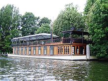 A large, low-slung boat lies moored on the bank of a body of water.  The lower deck is made from wood, interrupted by several large windows.  The upper deck is made from decorative metal, and covered with a large glass awning.