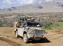A camouflaged military vehicle on top of a hill. The valley behind the vehicle contains several complexes of buildings and flowering trees. A steep mountain range is in the background