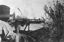 A soldier lies amongst the grass in the prone position behind a machine gun which he is holding in the shoulder