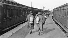 Soldiers walk down a station platform, while on either side of the platform are two trains. The men are wearing slouch hats and are carrying bags over their shoulders