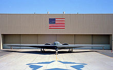 Front view of tailless aircraft parked in front of building. On the building face is a blue and red rectangular flag. In the foreground is a star shape on the ground