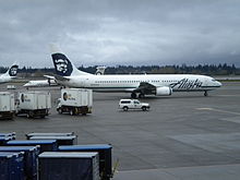 Right side view of an airplane taxiing on the tarmac, with several trucks in the foreground and to the left.  In the background is a tree-covered hill and dark clouds.
