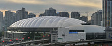 Cars pass by on an elevated highway in the foreground. In the midground is a large, oval-shaped building with a white, domed roof.
