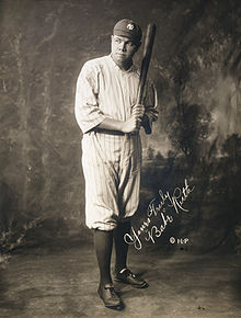 A man in full baseball attire wears a pinstriped jersey and a hat with overlapping white "N" and "Y". Looking to the left of the camera, he is holding a baseball upward.