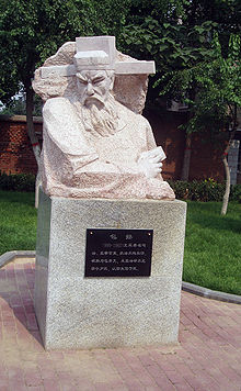 An marble statue depicting the upper half of an elderly man's body. The man has a long beard and thick eyebrows, and is wearing a square cut hat with long, thick, hoizontal protrusions coming out from the sides, near the ears. The carving is angular, and the figure being depicted appears to be in the middle of a sharp turn.