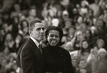 Barack and Michelle Obama, wearing dark outdoor clothes, in front of a crowd. His expression is muted; she has a wide smile.