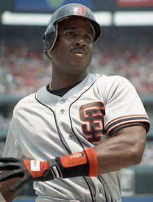 An African-American man looks just right of the camera. His helmet and white jersey both have an orange "S" over "F" logo on them. The man's left arm is crossed over his body and his right is out of the picture. There is a black and orange glove on his left hand.
