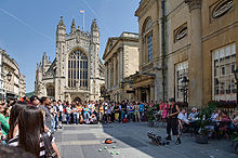 Gray paved area with lots of people around brightly dressed performer. To the right is a yellow stone building and in the background the tower of the abbey.