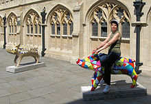 Two pig statues in front of yellow stone building with arched windows. The nearest status is multi coloured band being ridden by person.
