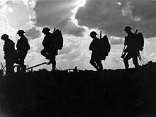 Five soldiers silhouetted against the sky. Rays of sun burst through dark clouds to create a dramatic and atmospheric shot. They are all wearing steel helmets, and three of them are clearly carrying rifles and backpacks.
