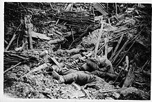 In the foreground the limp bodies of dead German soldiers lie amidst the rubble of a destroyed trench. It is difficult to distinguish the soldiers from the rubble around them but three bodies are visible. One man wearing a helmet, has been pushed forward by the blast and although dead, appears to crouch forward.