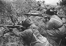A submachine-gunner and a machine-gunner lying in a trench with their weapons ready and pointing toward the left. In the background, several more soldiers are also lying in the trench and facing left.